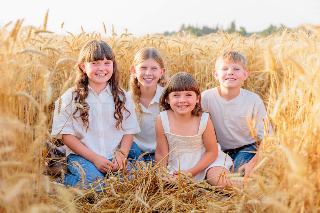 Four siblings smiling together while sitting in a wheat field at sunset.